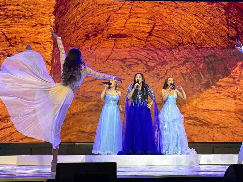 Three singers in gowns against a red-rock cave backdrop